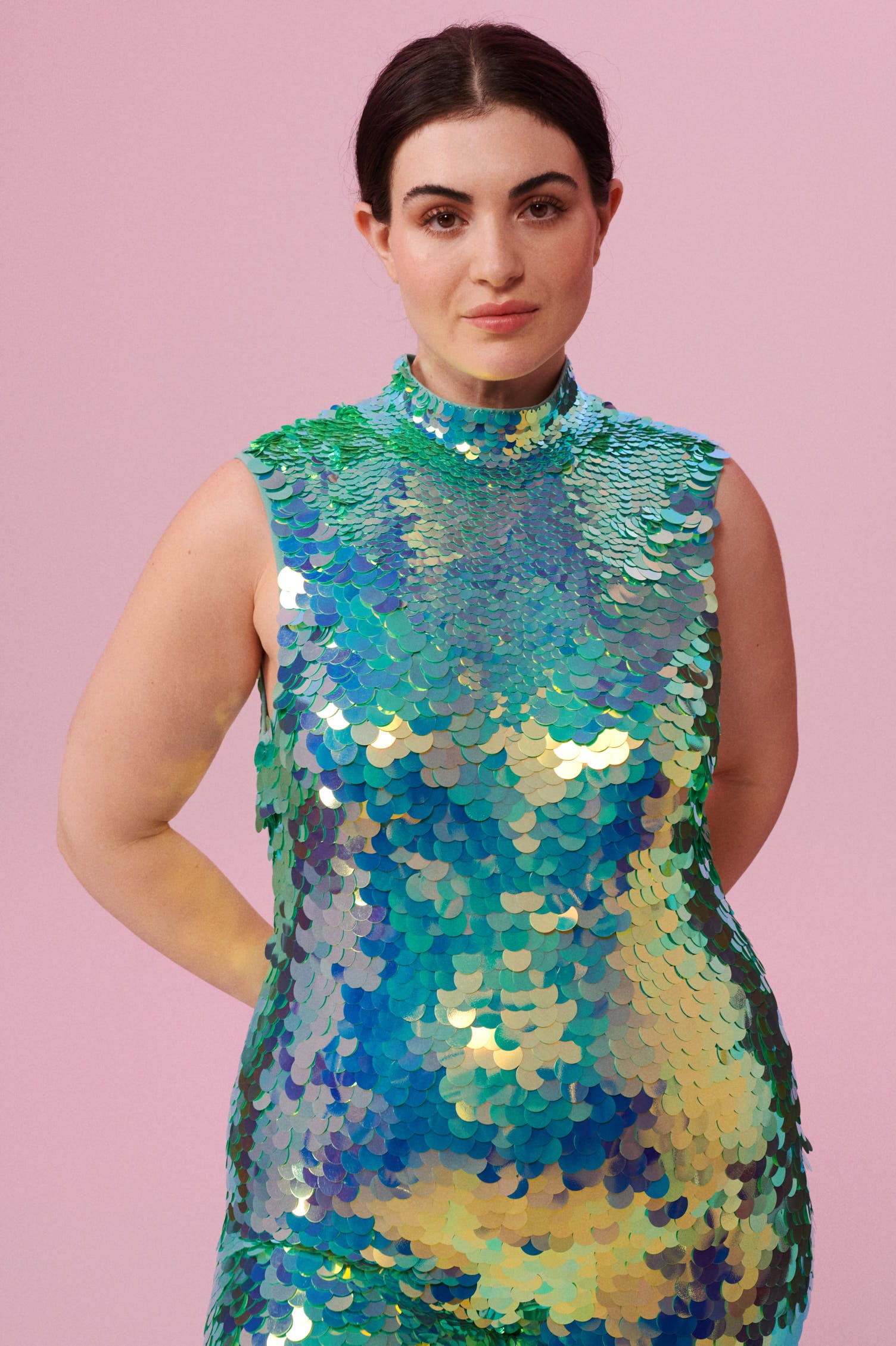 A woman with brown hair, wearing an all-in-one sequin stretchy festival jumpsuit made with large round holographic Rosa Bloom sequins. The Iris jumpsuit sequins glisten all over in this chameleon colour way, creating a mix of shimmering colours of soft mint green and sage that sparkles in the light.