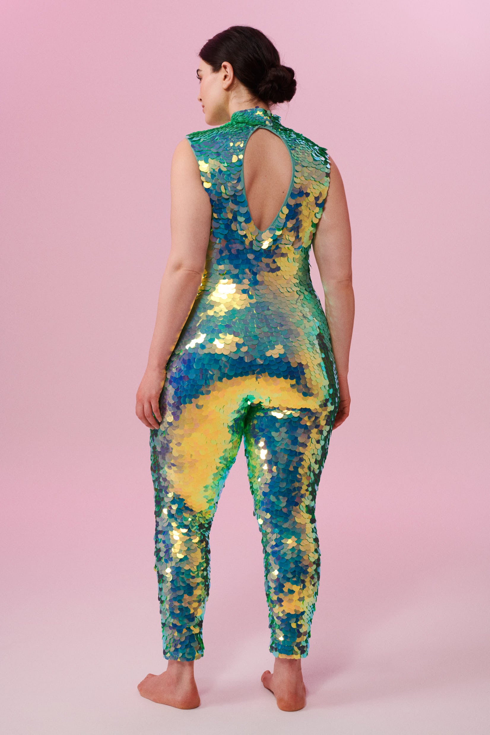 A woman with brown hair standing with her back to the viewer, wearing an all-in-one sequin stretchy festival jumpsuit made with large round holographic Rosa Bloom sequins. The Iris jumpsuit sequins glisten all over in this chameleon colour way, creating a mix of shimmering colours of soft mint green and sage that sparkles in the light.