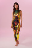 A woman with brown hair, wearing an all-in-one sequin stretchy festival jumpsuit made with large round holographic Rosa Bloom sequins. The woman  is wearing the Ember jumpsuit in front of a pink background. The sequins glisten all over in this ember colour way, creating a mix of shimmering colours of soft red, gold and yellow that sparkles in the light.