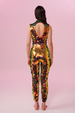A woman with brown hair, standing with her back to the viewer wearing an all-in-one sequin stretchy festival jumpsuit made with large round holographic Rosa Bloom sequins. The woman  is wearing the Ember jumpsuit in front of a pink background. The sequins glisten all over in this ember colour way, creating a mix of shimmering sequin colours of soft red, gold and yellow that sparkles in the light.