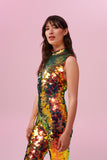 A woman with brown hair, wearing an all-in-one sequin stretchy festival jumpsuit made with large round holographic Rosa Bloom sequins. The woman  is wearing the Ember jumpsuit in front of a pink background. The sequins glisten all over in this ember colour way, creating a mix of shimmering colours of soft red, gold and yellow that sparkles in the light.