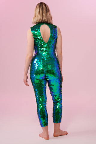 A woman with short blonde hair, standing with her back to the viewer wearing an all-in-one sequin stretchy festival jumpsuit made with large round holographic Rosa Bloom sequins. The woman  is wearing the Emerald jumpsuit in front of a pink background. The sequins glisten all over in this emerald colour way, creating a mix of shimmering sequin colours of green and blue that sparkles in the light.