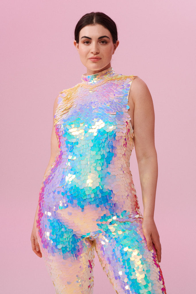 A woman with brown hair, standing facing the viewer wearing an all-in-one sequin stretchy festival jumpsuit with a high neck, made with large round holographic Rosa Bloom sequins. The woman  is wearing the Opal jumpsuit in front of a pink background. The sequins glisten all over in this Opal colour way, creating a mix of shimmering sequin colours of pinks, purples and blues that sparkles in the light.