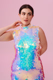 A crop of a woman with brown hair, standing facing the viewer wearing an all-in-one sequin stretchy festival jumpsuit with a high neck, made with large round holographic Rosa Bloom sequins. The woman  is wearing the Opal jumpsuit in front of a pink background. The sequins glisten all over in this Opal colour way, creating a mix of shimmering sequin colours of pinks, purples and blues that sparkles in the light.