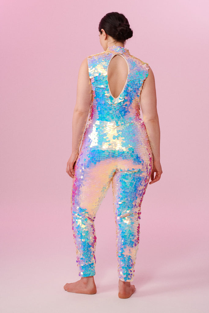 A rear view of a woman with brown hair, standing facing the viewer wearing an all-in-one sequin stretchy festival jumpsuit with a high neck, made with large round holographic Rosa Bloom sequins. The woman  is wearing the Opal jumpsuit in front of a pink background. The sequins glisten all over in this Opal colour way, creating a mix of shimmering sequin colours of pinks, purples and blues that sparkles in the light.