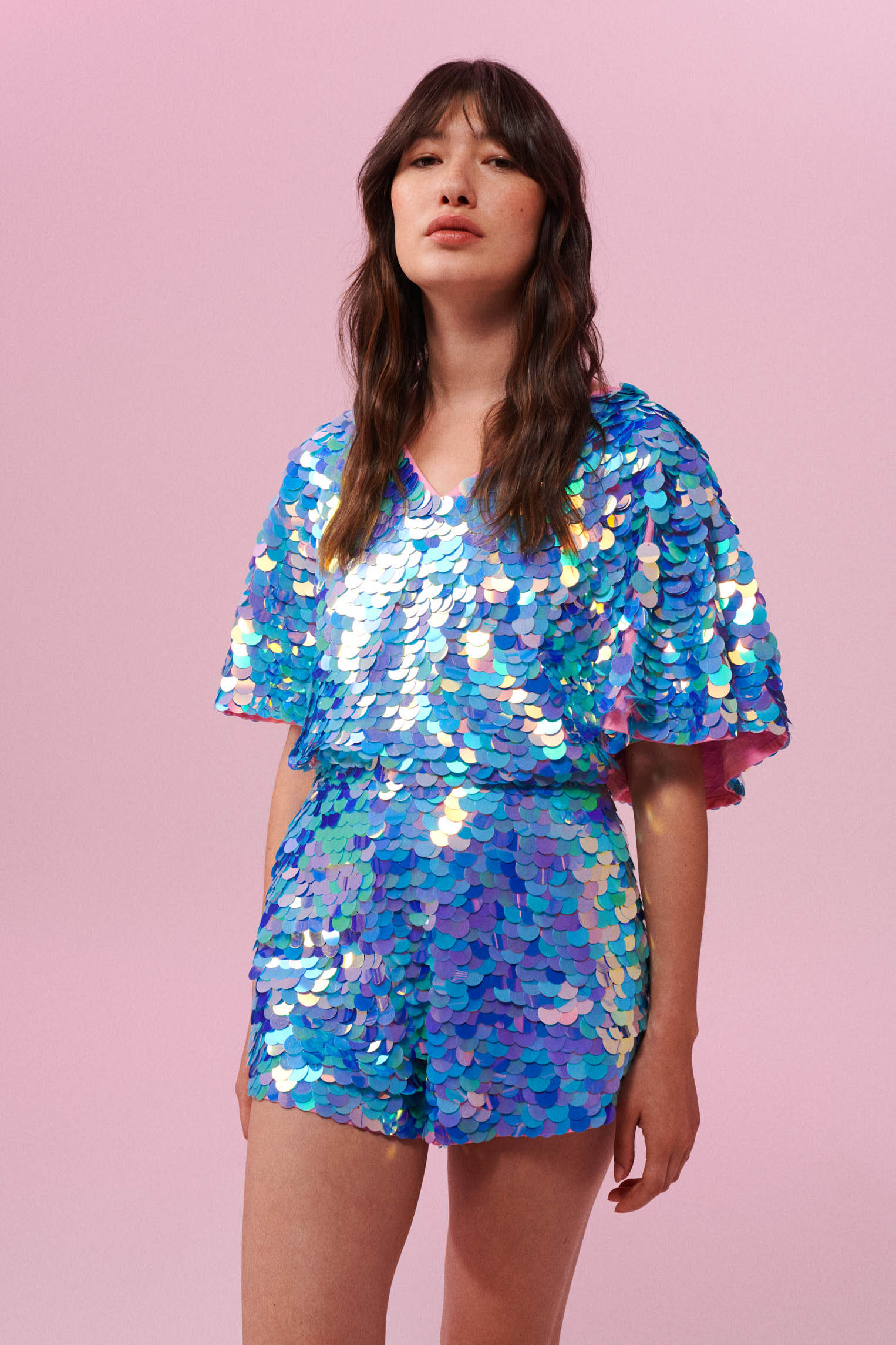 Dark haired model with long brown hair, wearing a Rosa Bloom sequin Mella romper playsuit in Amethyst design. Pink, blue and lilac sequins shimmer in the light showing the shorts and commanding cape sleeves, and acres of iridescent disc sequins. 
