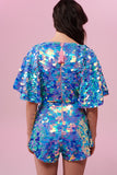 Dark haired model with her back to the camera wearing a Rosa Bloom sequin Mella romper in Amethyst design. Pink, blue and lilac sequins shimmer in the light showing the commanding cape sleeves and acres of iridescent disc sequins. 