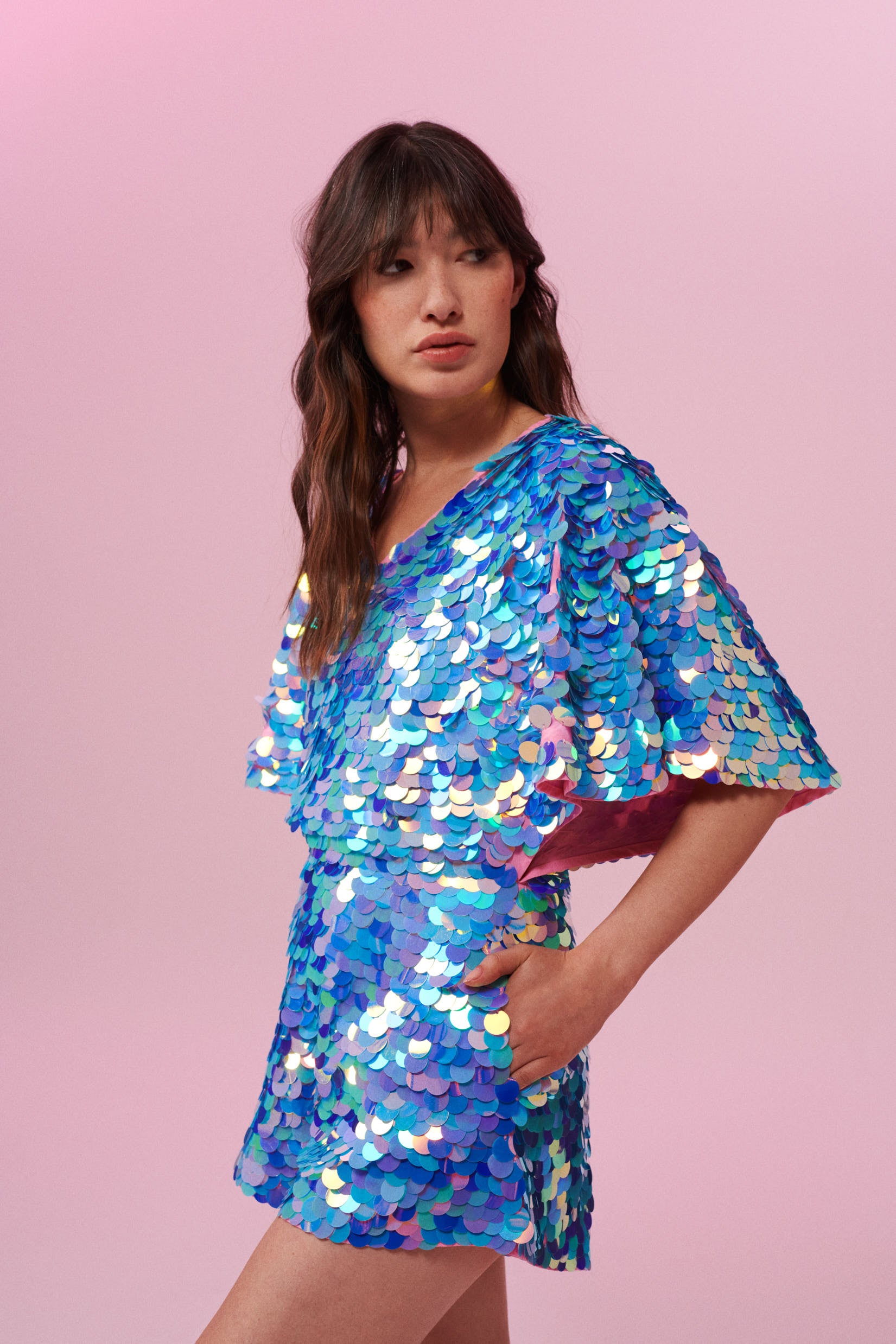 Dark haired model with long brown hair swept to the side, wearing a Rosa Bloom sequin Mella romper in Amethyst design. Pink, blue and lilac sequins shimmer in the light showing the commanding cape sleeves and acres of iridescent disc sequins. 