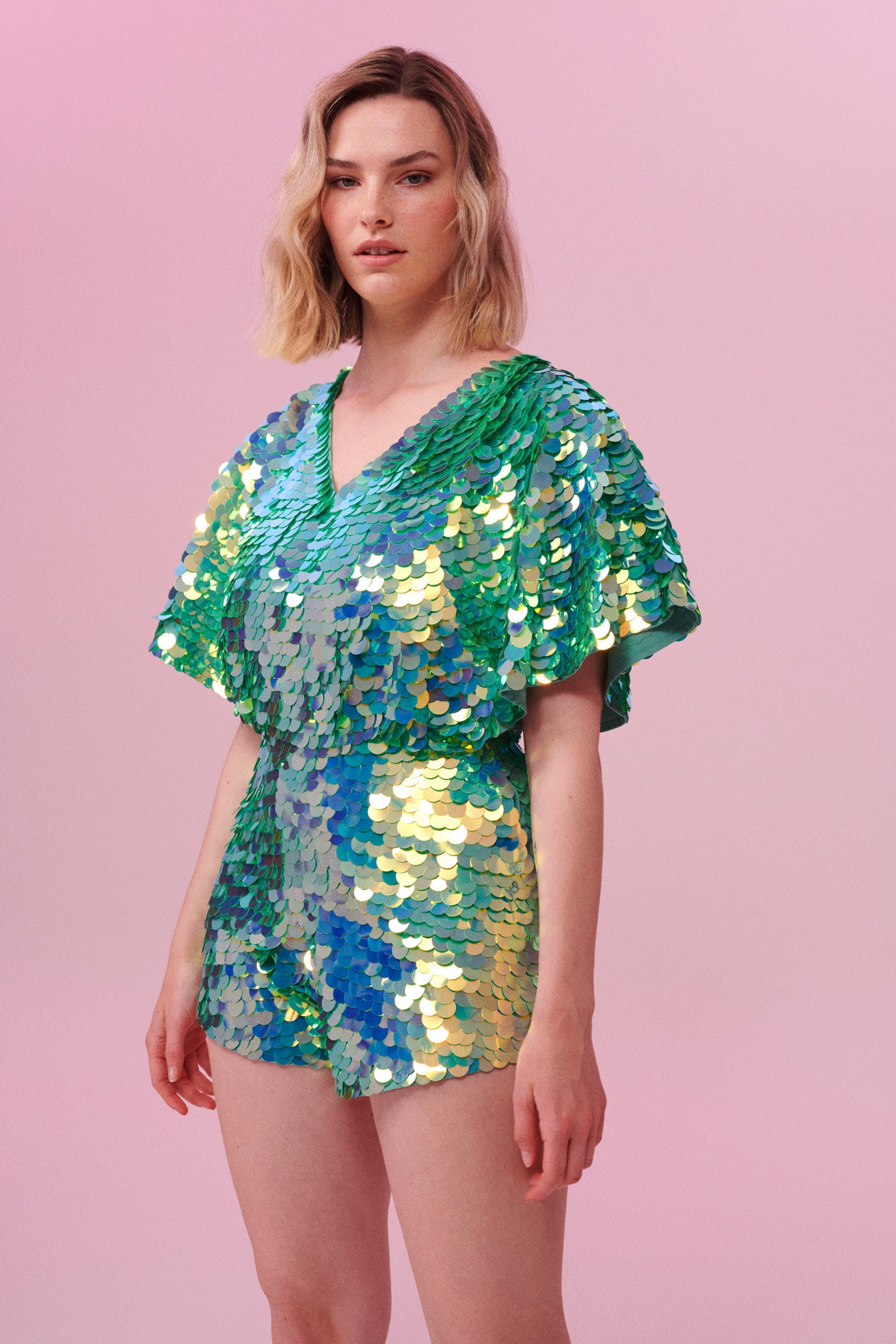 A model with short blonde hair wearing a Rosa Bloom sequin Mella romper playsuit in the chameleon design. A mix of green sequins shimmer in the light showing the shorts and commanding cape sleeved outfit.  The iridescent disc sequins, as worn by Taylor Swift shimmers in the light.  