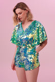 A model with short blonde hair wearing a Rosa Bloom sequin Mella romper playsuit in the chameleon design. A mix of green sequins shimmer in the light showing the shorts and commanding cape sleeved outfit.  The iridescent disc sequins, as worn by Taylor Swift shimmers in the light.  