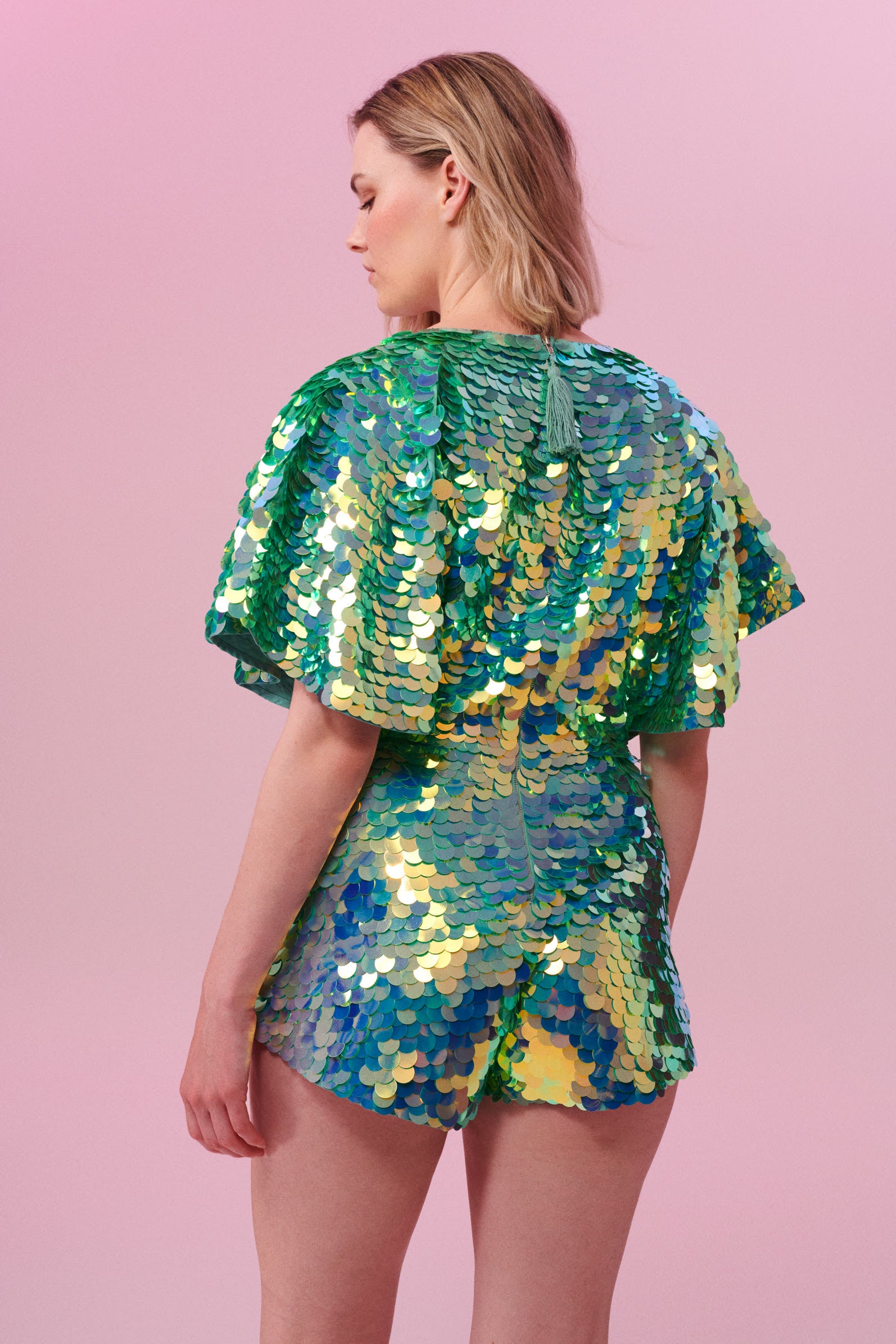 A model with short blonde hair with her back to the camera wearing a Rosa Bloom sequin Mella romper playsuit in the chameleon design. A mix of green sequins shimmer in the light showing the shorts and commanding cape sleeved festival outfit.  The all over iridescent disc sequins, as worn by Taylor Swift shimmers in the light.  