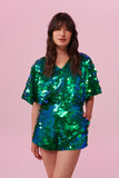 Dark haired model with long brown hair on a pink backdrop, wearing a Rosa Bloom sequin Mella romper playsuit in Emerald design. Green and blue, sequins shimmer in the light showing the shorts and commanding cape sleeves, and acres of iridescent disc sequins. 