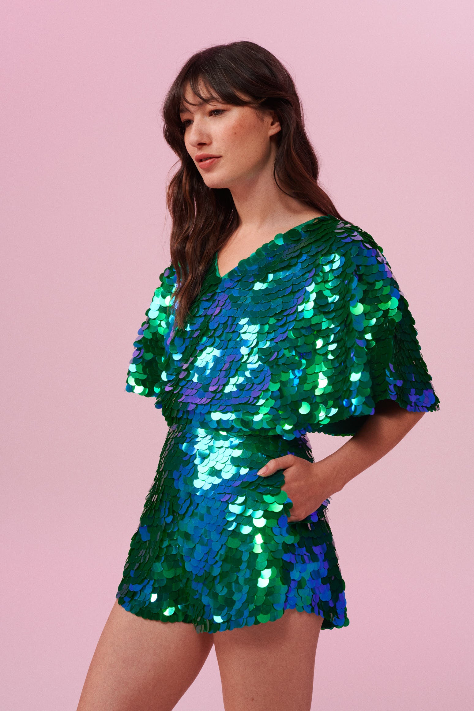 Dark haired model with long brown hair on a pink backdrop, wearing a Rosa Bloom sequin Mella romper playsuit in Emerald design. Green and blue, sequins shimmer in the light showing the shorts and commanding cape sleeves, and acres of iridescent disc sequins. 