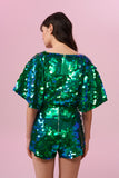 Dark haired model with long brown hair on a pink backdrop, with her back to the camera. She is wearing a Rosa Bloom sequin Mella romper playsuit in Emerald design. Green and blue, sequins shimmer in the light showing the shorts and commanding cape sleeves, and acres of iridescent disc sequins. 