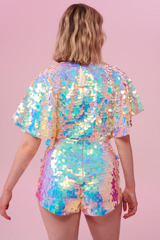 A model with short blonde hair with her back to the camera wearing a Rosa Bloom sequin Mella romper playsuit in the Opal design. A mix of white, pinks and blue sequins shimmer in the light showing the shorts and commanding cape sleeved festival outfit.  The all over iridescent disc sequins, as worn by Taylor Swift shimmers in the light.  