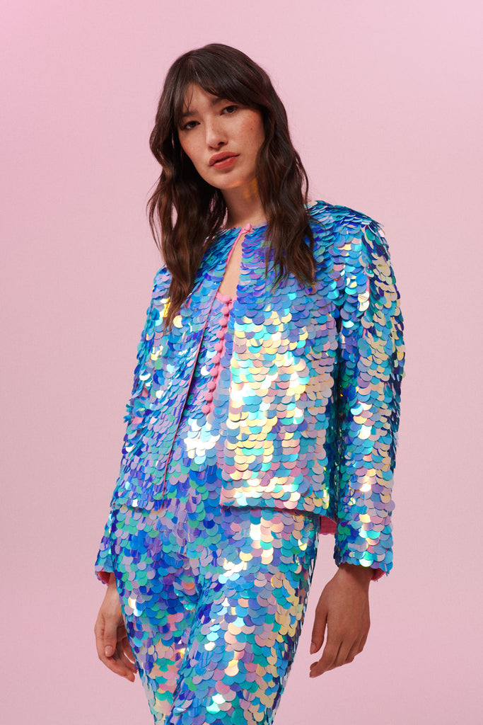 A woman with long brown hair standing front on to the camera in a brightly lit indoor room with pink walls wearing a Rosa Bloom blue and pink sequin jacket and Rosa Bloom blue and pink sequin jumpsuit