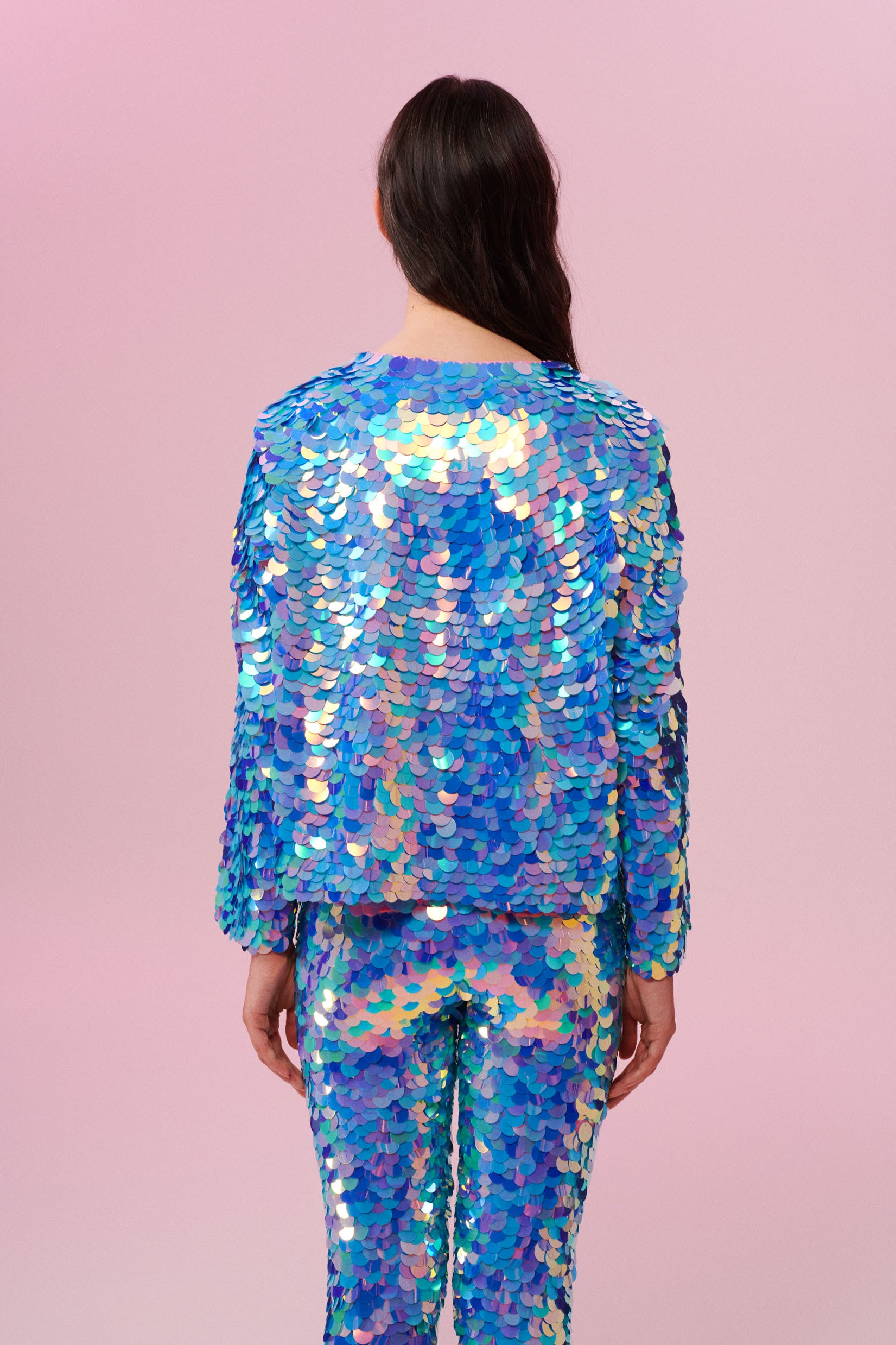 A woman with long brown hair swept over her shoulder standing with her back to the camera in a brightly lit indoor room with pink walls wearing a Rosa Bloom blue and pink sequin jacket and Rosa Bloom blue and pink sequin leggings