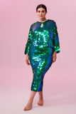 A plus size brunette model with her hair in a low bun wearing a Rosa Bloom Emerald green sequin dress and boxy sequin jacket
