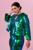 A plus size brunette model with her hair in a low bun and eyes lowered posing with her hand behind her head side on wearing a Rosa Bloom Emerald green sequin dress and boxy sequin jacket