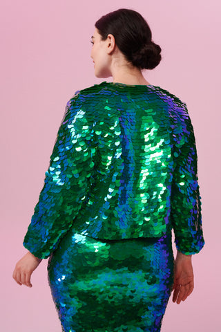 A plus size brunette model with her hair in a low bun posing with her back to the camera wearing a Rosa Bloom Emerald green sequin dress and boxy sequin jacket