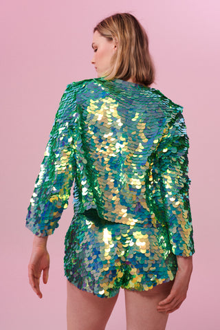 Blonde model with lob hair cut posing with her back to the camera wearing a full sequin outfit of Rosa Bloom green sequins, including a boxy Chanel-style jacket with a button top, a sequin crop top and sequin shorts