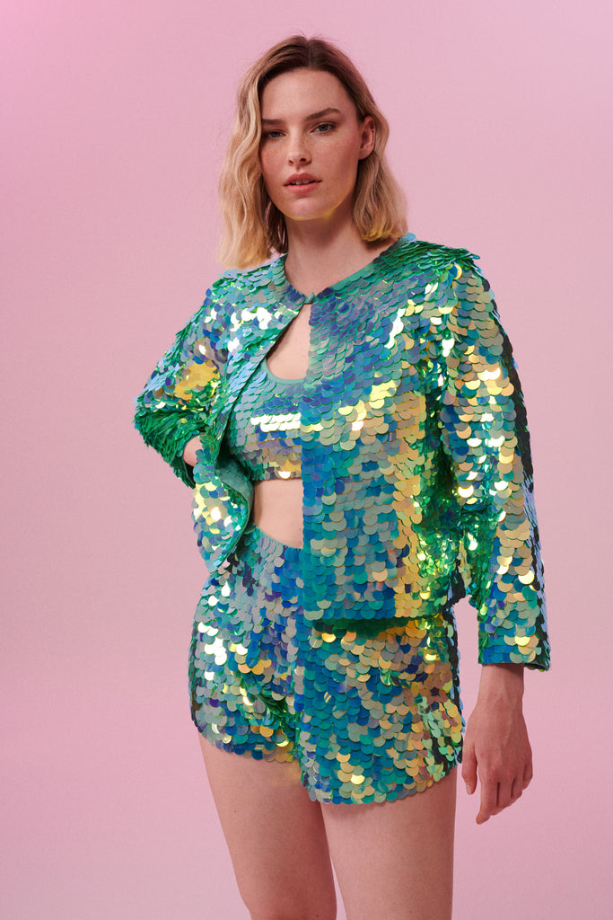 Blonde model with lob hair cut posing front on with her arm on her hip wearing a full outfit of Rosa Bloom green sequins, including a boxy Chanel-style jacket with a button top, a sequin crop top and sequin shorts