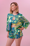 Blonde model with lob hair cut posing front on wearing a full outfit of Rosa Bloom green sequins, including a boxy Chanel-style jacket with a button top, a sequin crop top and sequin shorts