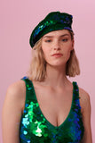 A woman with short blonde hair, wearing a sequin beret made from small round holographic sequins with hues of green and blue. The festival sequin hat matches the sequin jumpsuit that the model is wearing. The Emerald sequins by Rosa Bloom glisten, creating a mix of shimmering colours of emerald green and ultramarine blue.