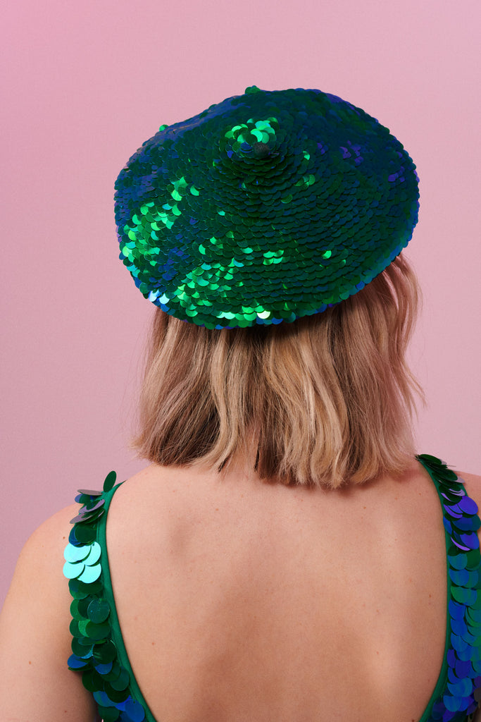 Rear view of a woman wearing a sequin beret made from small round holographic sequins with hues of green and blue. The festival sequin hat matches the sequin jumpsuit that the model is wearing. The Emerald sequins by Rosa Bloom glisten, creating a mix of shimmering colours of emerald green and ultramarine blue. 