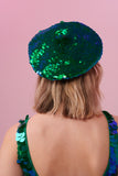 Rear view of a woman wearing a sequin beret made from small round holographic sequins with hues of green and blue. The festival sequin hat matches the sequin jumpsuit that the model is wearing. The Emerald sequins by Rosa Bloom glisten, creating a mix of shimmering colours of emerald green and ultramarine blue. 