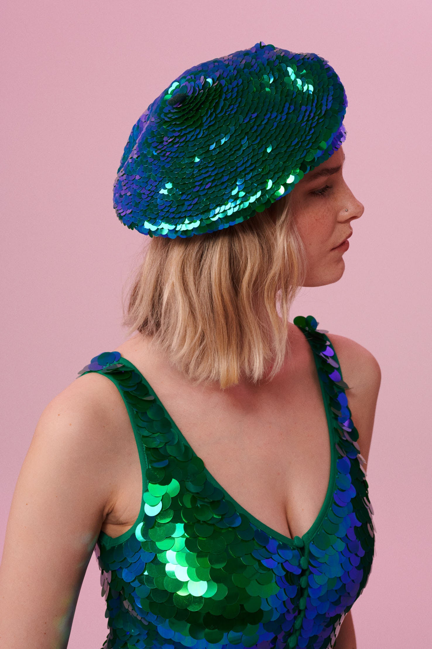 A woman with short blonde hair, wearing a sequin beret made from small round holographic sequins with hues of green and blue. The festival, woollen and sequin hat matches the sequin jumpsuit that the model is wearing. The Emerald sequins by Rosa Bloom glisten, creating a mix of shimmering colours of emerald green and ultramarine blue. 