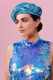A woman with brown hair tied back in a bun, facing to the side and wearing an iridescent blue, lilac and pink sequin festival beret. She is also wearing a high necked sequin top that matched the sequin hat with large and small sequins. The Amethyst sequins glisten, creating a mix of shimmering colours.