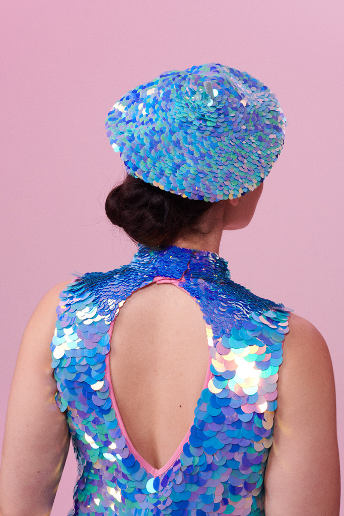 Rear view of a woman with brown hair tied back in a bun,  wearing an iridescent blue, lilac and pink sequin festival beret. She is also wearing a high necked sequin top with a cut-out back section that matches the sequin hat with large and small sequins. The Amethyst sequins glisten, creating a mix of shimmering colours.