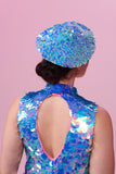 Rear view of a woman with brown hair tied back in a bun,  wearing an iridescent blue, lilac and pink sequin festival beret. She is also wearing a high necked sequin top with a cut-out back section that matches the sequin hat with large and small sequins. The Amethyst sequins glisten, creating a mix of shimmering colours.