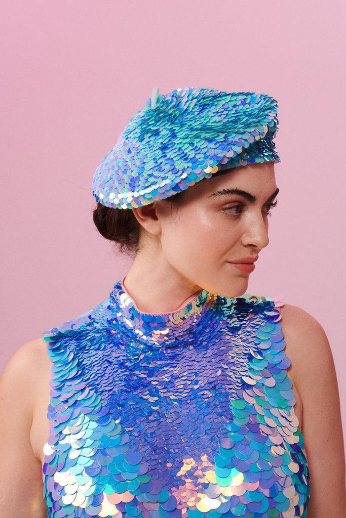 A woman with brown hair tied back in a bun, facing to the side and wearing an iridescent blue, lilac and pink sequin festival beret. She is also wearing a high necked sequin top that matched the sequin hat with large and small sequins. The Amethyst sequins glisten, creating a mix of shimmering colours.