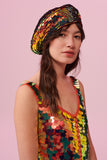 A woman facing to the side, with brown hair swept to the side, wearing a sequin beret made from small round holographic sequins with hues of gold, red, burnt orange and yellow. The festival, woollen and sequin hat matches the sequin jumpsuit that the model is wearing. The Fox sequins by Rosa Bloom glisten, creating a mix of shimmering colours.