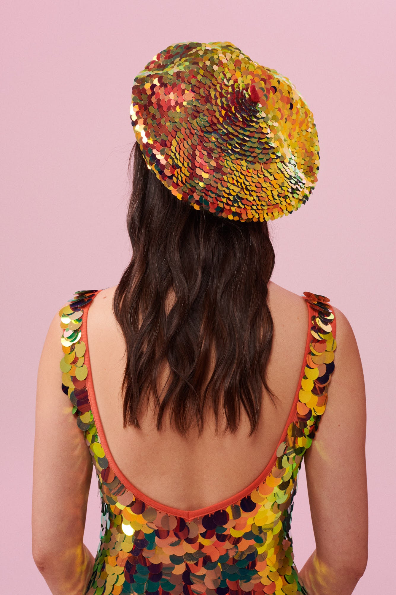  Rear of a woman with brown, wearing a sequin beret made from small round holographic sequins with hues of gold, red, burnt orange and yellow. The festival, woollen and sequin hat matches the sequin jumpsuit that the model is wearing. The Fox sequins by Rosa Bloom glisten, creating a mix of shimmering colours.