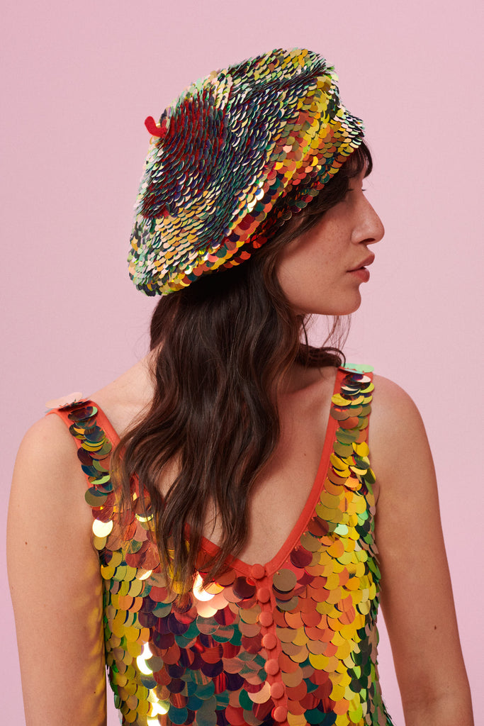A woman facing to the side, with brown hair swept to the side, wearing a sequin beret made from small round holographic sequins with hues of gold, red, burnt orange and yellow. The festival, woollen and sequin hat matches the sequin jumpsuit that the model is wearing. The Fox sequins by Rosa Bloom glisten, creating a mix of shimmering colours.
