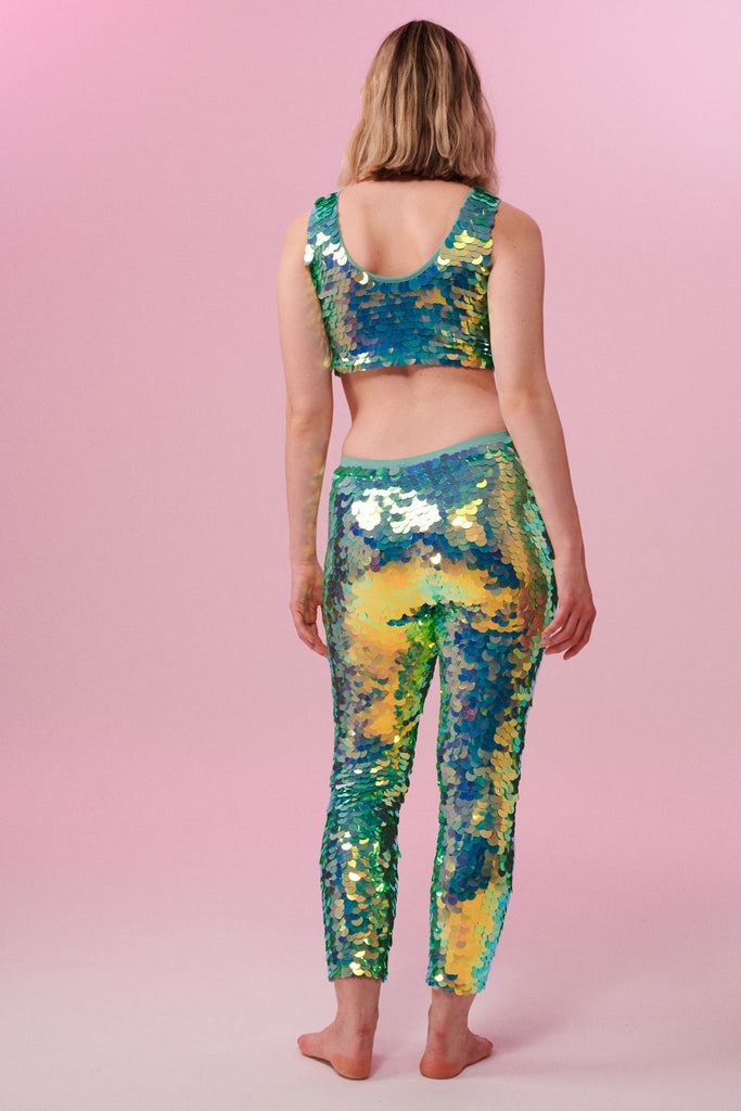 A rear view of a woman wearing iridescent mint green stretchy sequin Indus leggings covered in large round holographic Rosa Bloom sequins. The sequins glisten, creating a mix of shimmering colours of pale green sequinned mint and warm peachy gold make this sequin glow. The model is also wearing a matching stretchy sequin vest top, in matching colours. This Chameleon sequin outfit looks like it is glowing. 
