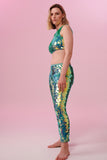 A woman wearing iridescent mint green stretchy sequin Indus leggings covered in large round holographic Rosa Bloom sequins. The sequins glisten, creating a mix of shimmering colours of pale green sequinned mint and warm peachy gold make this sequin glow. The model is also wearing a matching stretchy sequin vest top, in matching colours. This Chameleon sequin outfit looks like it is glowing. 