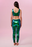 A rear view of a woman wearing iridescent blue and green Indus leggings covered in large round holographic Rosa Bloom sequins. The sequins glisten, creating a mix of shimmering colours make this sequin glow. The model is also wearing a matching stretchy sequin vest top, in matching colours. This Emerald sequin outfit looks like it is glowing. 