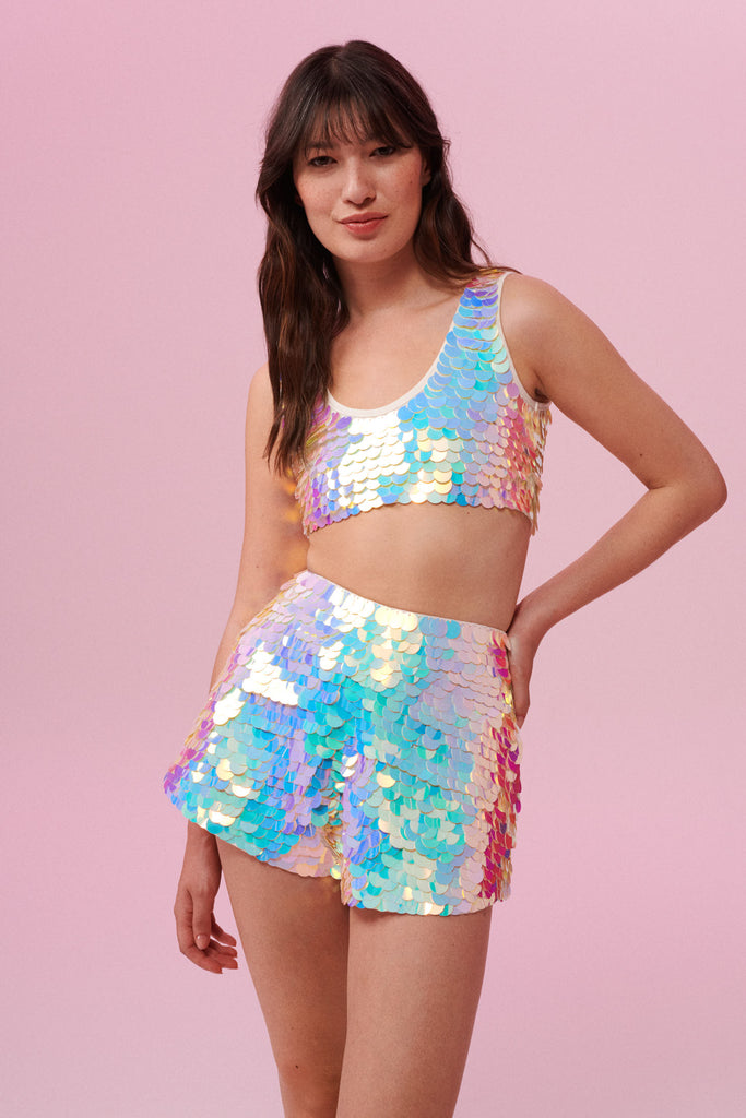 A woman wearing a festival sequin vest and matching shorts. The Juno high waisted, tailored sequin shorts are covered in large round holographic Rosa Bloom sequins. The sequins glisten, creating a mix of shimmering colours in  iridescent purple, pink white and blue. The model is also wearing a matching stretchy sequin cropped vest, in the same coloured sequins. 
