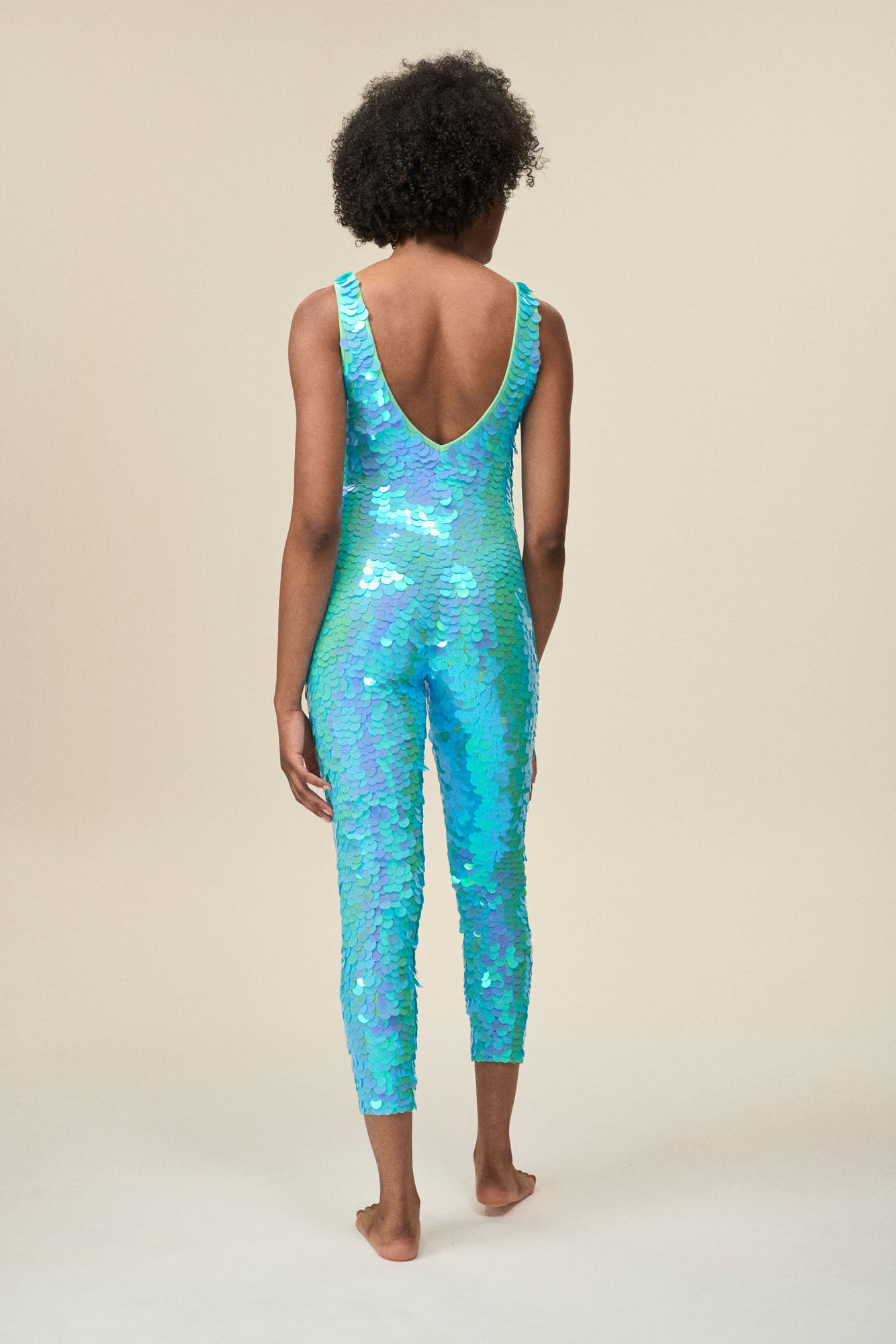 Back view of a woman wearing the Rosa Bloom Electra jumpsuit in a mesmerising aqua blue. A stretchy jumpsuit with low V back detail that hugs and contours her body embellishing her in sparkling sequins.