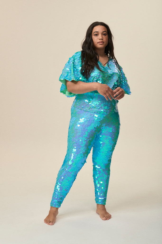 A woman facing forward wearing a mint sequin top with large cape sleeves covered in large round holographic aqua blue coloured sequins.