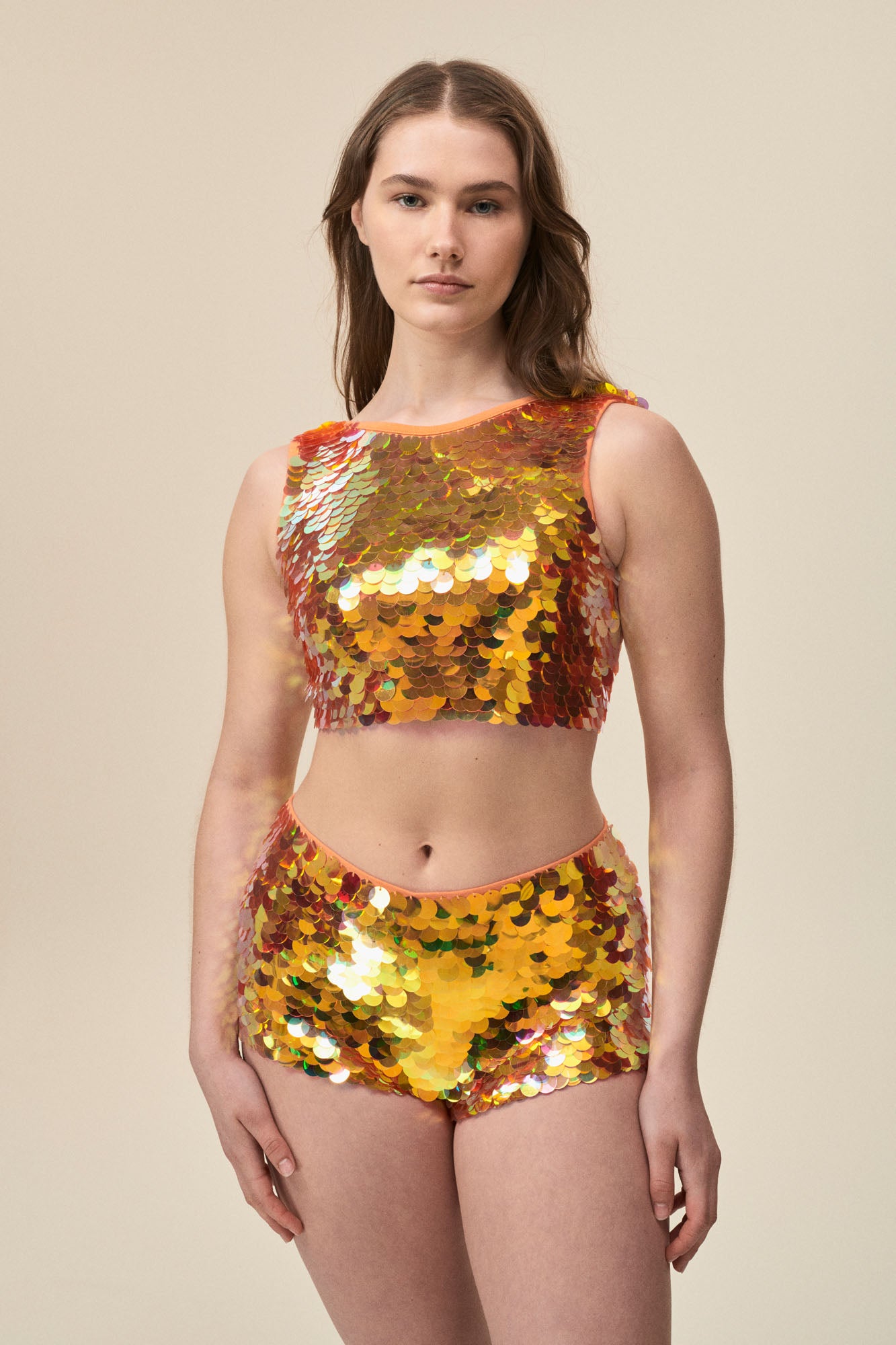 A woman faces you wearing stretchy Rosa Bloom Gigi hotpants with large sparkling orange sequins