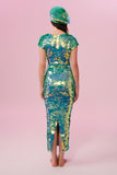 A woman standing with her back to the viewer wearing an iridescent pale green sequin long dress. A figure hugging  dress down to the ankles with small capped sleeves completely covered in large round holographic Rosa Bloom sequins. The sequin colours shine in the studio lights, creating a mix of shimmering colours of sage green, mint and a warm peachy gold making this sequin dress glow when worn. The model is also wearing a matching sequinned beret hat, slightly to the side of her head. 