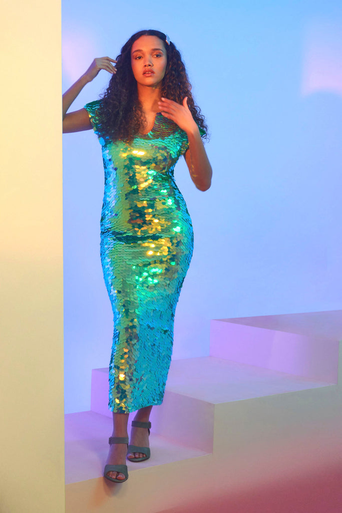 A woman with long brown curls hair, facing the viewer wearing an iridescent pale green sequin long dress. A figure hugging  dress down to the ankles with small capped sleeves completely covered in large round holographic Rosa Bloom sequins. The sequin colours shine in the studio lights, creating a mix of shimmering colours of sage green, mint and a warm peachy gold making this sequin dress glow when worn.