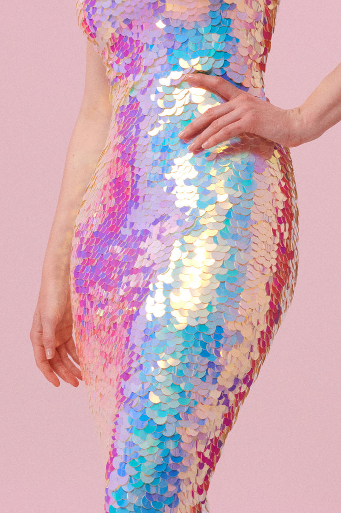 A crop of a figure with her hands on her hips, wearing a figure hugging long sequin dress completely covered in large round holographic Rosa Bloom sequins. The sequins glisten, creating a mix of shimmering colours of pink, blue, lilac and white.