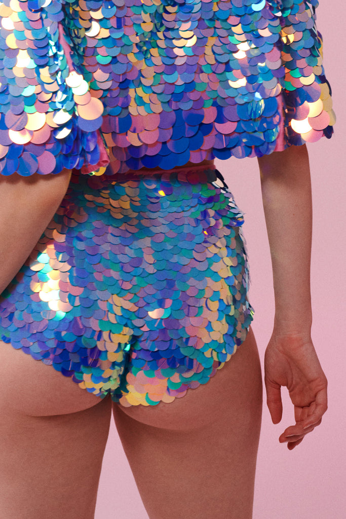A crop of the back of a figure, wearing high waisted shorts and a cape completely covered in large round holographic Rosa Bloom sequins. The sequins glisten in the light, creating a mix of shimmering colours of pink, blue and lilac. 