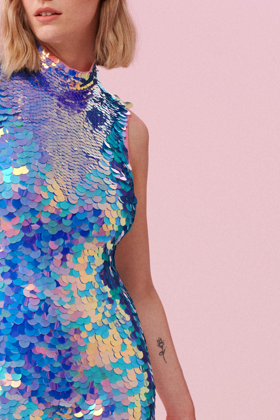 A crop of a woman with short blonde hair, wearing an all in one sequin stretchy festival jumpsuit made with large round holographic Rosa Bloom sequins. The Iris sequins glisten, creating a mix of shimmering colours of pink and blue.  The Amethyst sequins by Rosa Bloom glisten, creating a mix of shimmering colours.   